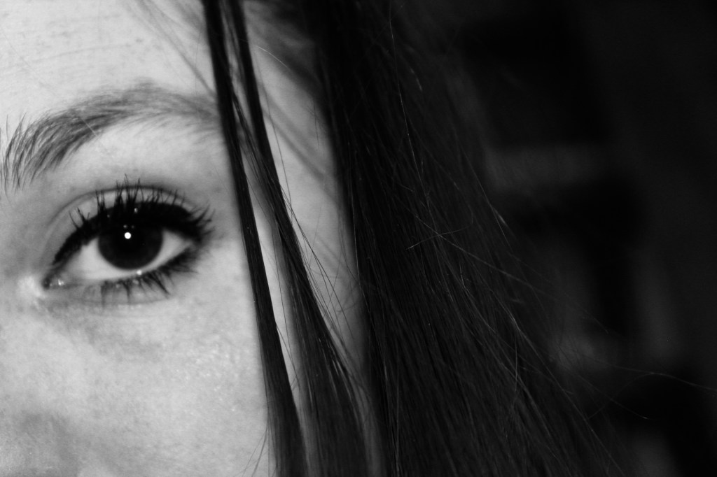 The frame is a close up of the woman's eye. 1/3 of the shot is her face, only the frame holds from her cheek bone to just above her eyebrow. A few strands of hair accompany it and the rest of the image is black, matching the color of the woman's eye. 