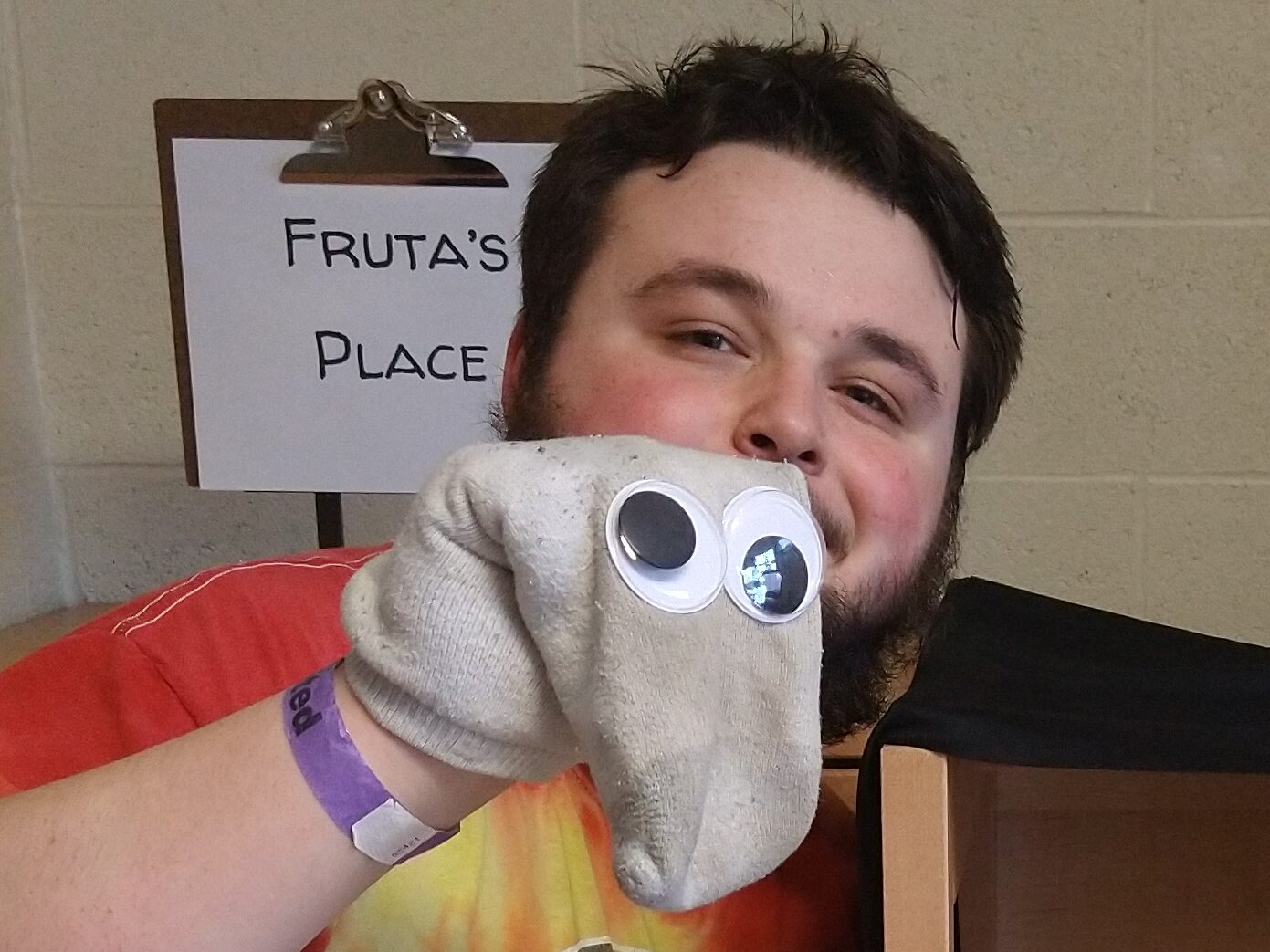 Dingy white sock with very large googley eyes, flopped over as if to have a long snout. The sock is in front of a sign that says "Fruta's Place".