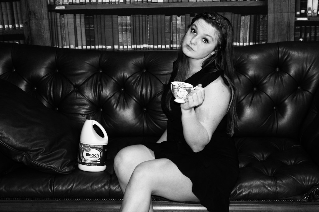 The woman sits on the couch, her knees bend on an angle with her body facing left. Her forward left elbow is bent nearly completely but keeping the tea cup in her hand straight. The bottle of bleach next her her knee. Her head is titled and there is no smile on her face. Guilt looms over her like a dark cloud. 