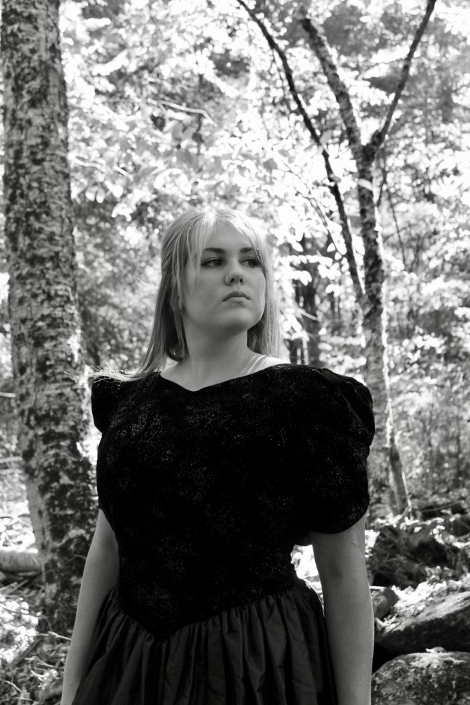 The photographer is positioned below the woman so the angle is looking up on her. She is on a slight left angle. Her shoulders are back, her chin is held high, her eyes look to the left. Her hair is behind her showing her strong facial expression. The woods blur behind her as she is the center focus of the frame. 