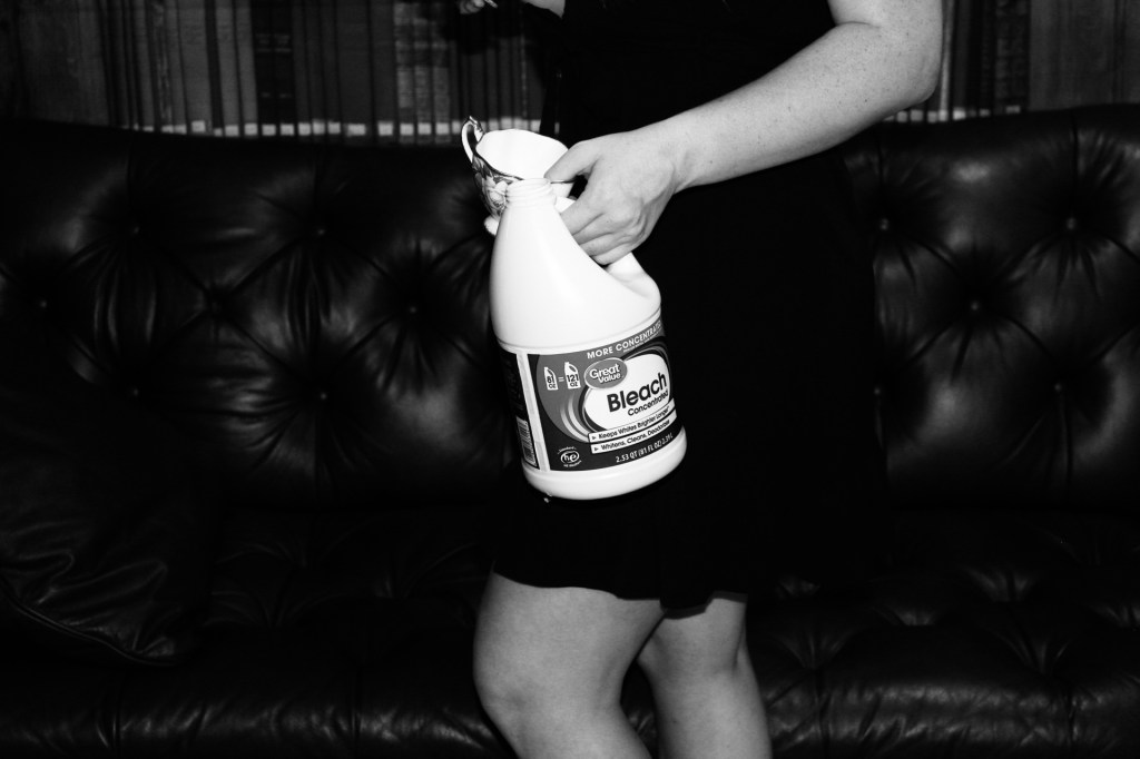 All that is visible is the woman lower torso down to her knees. She is standing in front of the couch on a side profile with her front knee bent mimicking her left forward arm. In her left hand she holds a bleach bottle and tea cup which are the focal point of the image. 