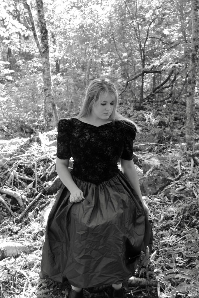 The woman sits in a chair unaccompanied by anything else except the woods surrounding her. Her right arm is slightly bent grasping her dress at the mid thigh, her left arm falls behind her. Shoulders straight, she looks down at an angle towards her left foot. The dress reaches the ground. The dress has a v shaped torso giving a subtle witch like look to her. 
