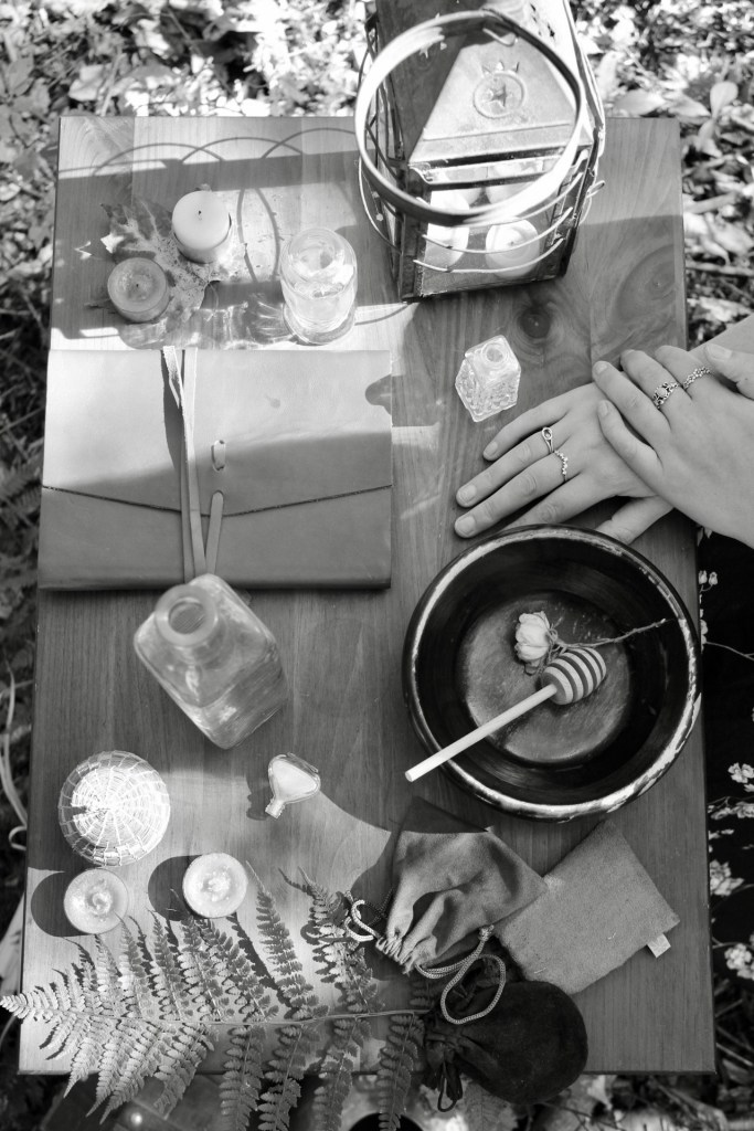 A wooden table covered in magical little trinkets. Three tea candles scattered on the edges, four vases varying in size and height, three plush bags, a lantern a leather bound notebook, a wooden bowl with a honey stick and a flower inside, all surrounding a pair of hands. These hands are overlapping one another and the woman wears rings on both her index & middle finger as well as her right ring finer and middle finger. 