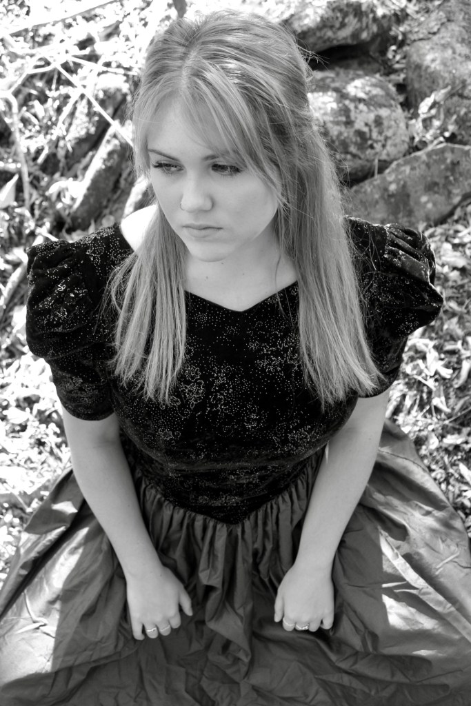 The woman sits with her legs spread and her hands on each inner thigh. The photo is captured from above her. The dress she is wearing makes her masculine stance softer and less noticeable. 