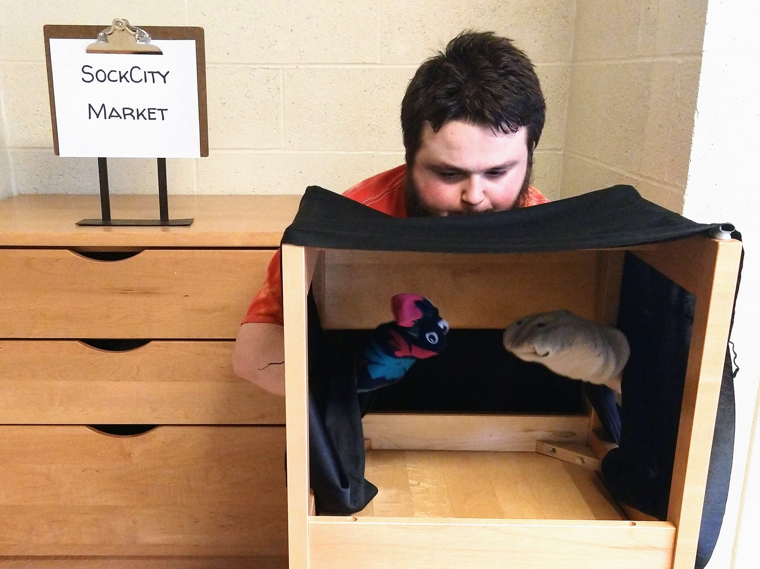 Man holds up Chausette and Hanes in the puppet theatre. The sign behind him says "SockCity Market"