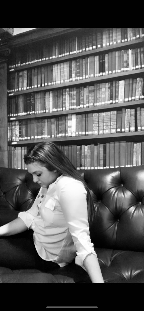 The woman sits on the couch with her head facing down. Her hair is pushed behind her. She is leaning on one arm. 